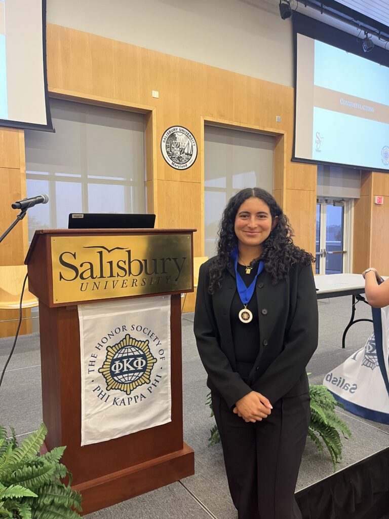 Gibelle Beaujon stands proudly in front of a podium at Salisbury University during the Phi Kappa Phi National Honor Society induction ceremony. She is dressed in formal black attire and wears a medal around her neck, symbolizing her induction. Behind her, a banner with the Phi Kappa Phi emblem hangs from the podium, and a projection screen in the background reads "Congratulations."